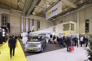 Messestand in einer großen Ausstellungshalle zum Thema Smart Living und E‑Haus, mit einem ausgestellten Elektrofahrzeug im Vordergrund und mehreren Besuchern, die sich informieren und austauschen.