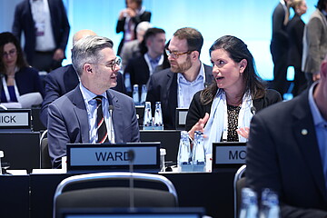 Mehrere Personen sitzen an Tischen in einem modern beleuchteten Konferenzsaal. Im Vordergrund unterhalten sich zwei Teilnehmende miteinander. Auf den Tischen stehen Wasserflaschen, Mikrofone und Namensschilder. Im Hintergrund sind weitere Teilnehmende zu sehen.