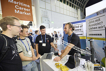 Besucherinnen und Besucher informieren sich an einem Unternehmensstand auf der SMM auf dem Maritime Career Market; auf dem Tresen sind ein Laptop und ein Modellkran zu sehen.