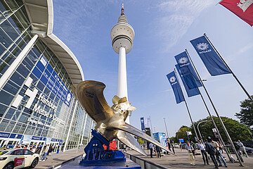 Blick auf das Messegelände Hamburg mit dem Heinrich-Hertz-Turm im Zentrum. Im Vordergrund steht eine große Schiffspropeller-Skulptur, rechts wehen mehrere blaue Messeflaggen. Vor dem Eingang der Hamburg Messe & Congress sind Besucherinnen und Besucher unterwegs unter blauem Himmel.