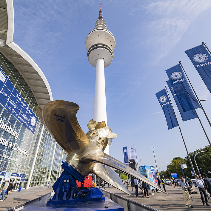 Blick auf das Messegelände Hamburg mit dem Heinrich-Hertz-Turm im Zentrum. Im Vordergrund steht eine große Schiffspropeller-Skulptur, rechts wehen mehrere blaue Messeflaggen. Vor dem Eingang der Hamburg Messe & Congress sind Besucherinnen und Besucher unterwegs unter blauem Himmel.