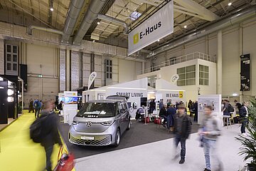 Messehalle mit einem Stand des „E-Haus“, dekoriert mit Bannern zu „Smart Living“. Im Vordergrund steht ein silberner VW ID. Buzz, mehrere Personen sind am Stand oder gehen vorbei.