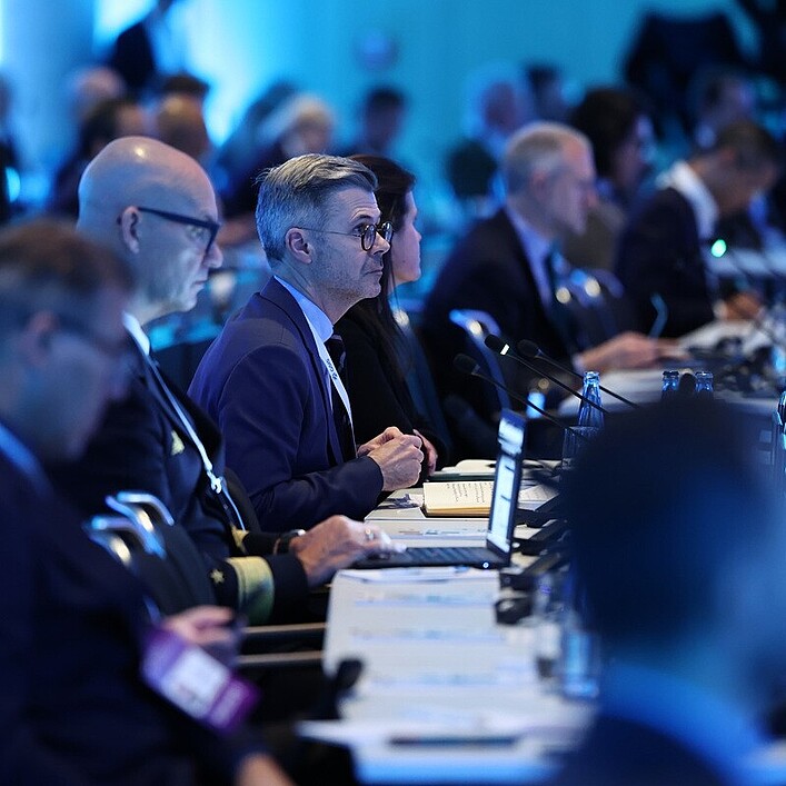 Reihe von Teilnehmenden bei einer Konferenz oder Sitzung, die an langen Tischen mit Mikrofonen und Laptops sitzen und aufmerksam nach vorne ausgerichtet sind; der Raum ist in kühles, blaues Licht getaucht, im Hintergrund sind weitere unscharfe Personen und Technik zu erkennen.