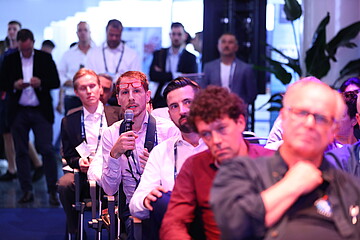 Eine Person sitzt bei einer Veranstaltung im Publikum und hält ein Mikrofon, während eine Frage gestellt oder ein Beitrag gemacht wird. Die Umgebung ist ein gut besuchter Konferenz- oder Networking-Bereich mit mehreren sitzenden und stehenden Teilnehmenden im Hintergrund, die aufmerksam zuhören.