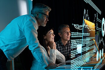 Drei Fachkräfte untersuchen gemeinsam ein technisches Gerät mit sichtbaren Leiterplatten in einer modernen Entwicklungs- oder Laborumgebung. Auf einem Schreibtisch stehen Computerbildschirme mit technischen Darstellungen, während eine Arbeitsleuchte den Versuchsaufbau beleuchtet. Im Hintergrund sind weitere technische Einrichtungen und ein Roboterarm zu erkennen.