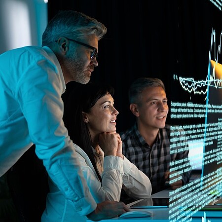 Drei Fachkräfte untersuchen gemeinsam ein technisches Gerät mit sichtbaren Leiterplatten in einer modernen Entwicklungs- oder Laborumgebung. Auf einem Schreibtisch stehen Computerbildschirme mit technischen Darstellungen, während eine Arbeitsleuchte den Versuchsaufbau beleuchtet. Im Hintergrund sind weitere technische Einrichtungen und ein Roboterarm zu erkennen.