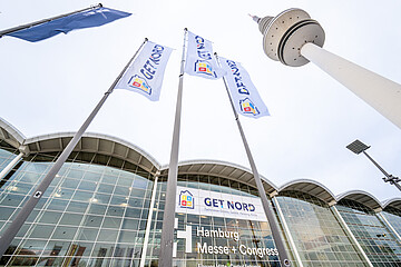 Blick von unten auf mehrere Fahnen mit der Aufschrift „GET NORD“, die vor dem Gebäude der Hamburg Messe + Congress im Wind wehen; im Hintergrund sind die Glasfassade, der Eingangsbereich mit Beschriftung sowie ein hoher Fernsehturm und eine Straßenleuchte zu sehen.