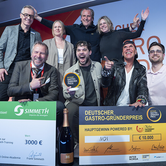 Gruppe von acht Personen auf einer Bühne bei einer Preisverleihung. Im Vordergrund werden ein Gewinnerpokal sowie zwei große Gewinnschecks des Deutschen Gastro-Gründerpreises präsentiert.