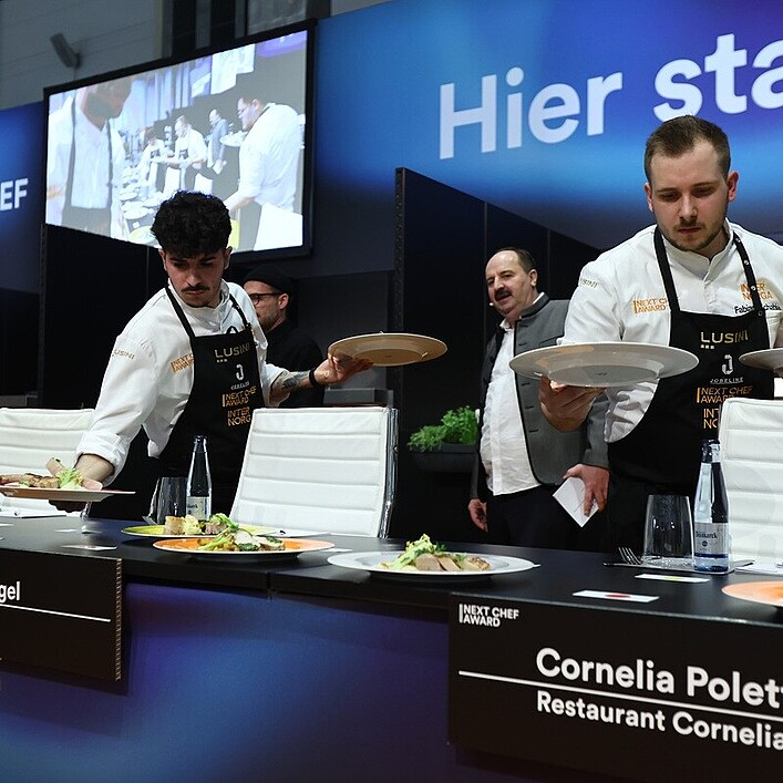 Mehrere Köche in weißer Arbeitskleidung servieren Teller mit angerichteten Speisen an einem Jurytisch beim „Next Chef Award“. Vor den weißen Stühlen stehen Namensschilder, unter anderem für Philipp Vogel („Orania Berlin“) und Cornelia Poletto („Restaurant Cornelia Poletto“). Im Hintergrund sind ein großer Bildschirm mit einer Live-Küchenaufnahme und die Worte „Hier starte…“ auf einer blauen Wand zu sehen.