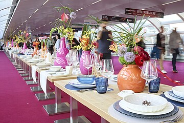 Ein sehr langer, festlich gedeckter Tisch steht in einer modernen, überdachten Passage mit pinkfarbenem Teppich. Der Tisch ist mit weißem Geschirr, blauen Gläsern und glänzendem Besteck eingedeckt. In der Tischmitte stehen zahlreiche farbenfrohe Vasen in Pink, Orange und weiteren Tönen, gefüllt mit üppigen Blumenarrangements aus verschiedenen Blüten und Gräsern. Im Hintergrund gehen mehrere Menschen an der Tafel vorbei, leicht verschwommen durch die Bewegung. Über der Szene hängen Schilder, auf denen unter anderem „Weltpremiere“ steht.