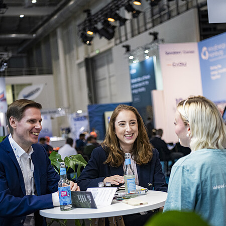 Drei Personen sitzen an einem Stehtisch auf einer Messe und führen ein Gespräch. Im Hintergrund sind Messestände, Besucherinnen und Besucher sowie Ausstellungsflächen in einer modernen Veranstaltungshalle zu sehen.