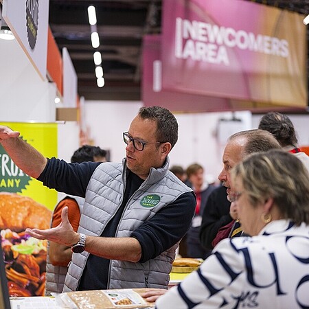 Eine Person auf einer Messe erklärt einer kleinen Gruppe von Besucherinnen und Besuchern ausführlich ein Produkt an einem Messestand. Im Hintergrund sind bunte Banner und Schilder zu sehen, darunter ein Bereich mit der Aufschrift Newcomers Area. Auf dem Tisch liegen verschiedene Lebensmittelproben und Informationsmaterialien.
