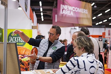 Eine Person auf einer Messe erklärt einer kleinen Gruppe von Besucherinnen und Besuchern ausführlich ein Produkt an einem Messestand. Im Hintergrund sind bunte Banner und Schilder zu sehen, darunter ein Bereich mit der Aufschrift Newcomers Area. Auf dem Tisch liegen verschiedene Lebensmittelproben und Informationsmaterialien.