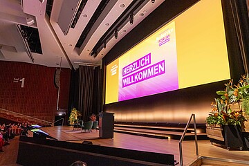 Großer Konferenzsaal mit Bühne und riesiger Leinwand. Auf dem Bildschirm steht „HERZLICH WILLKOMMEN“ vor gelb-pinkem Hintergrund des International Foodservice Forum. Die Bühne ist mit Sesseln, Rednerpult und tropischen Pflanzen dekoriert, während links Zuschauer im Auditorium sitzen.
