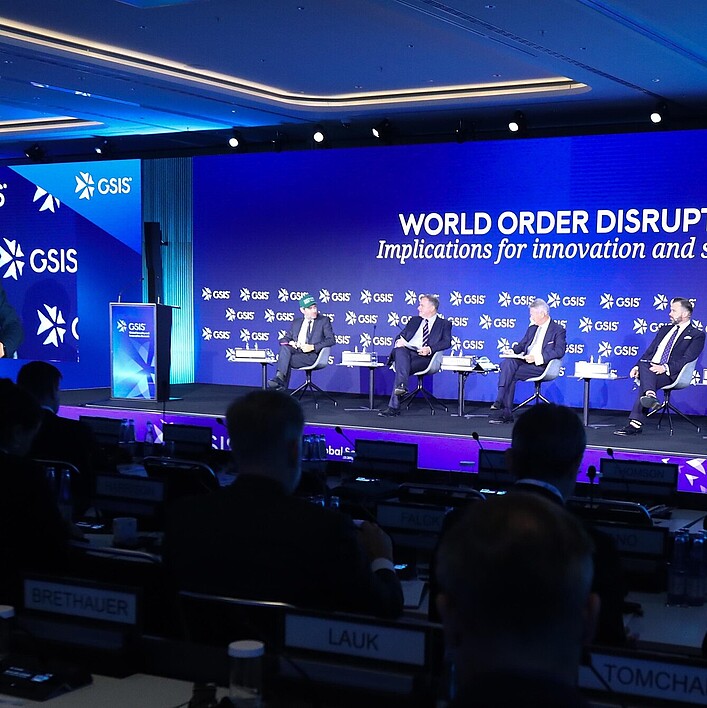 Podiumsdiskussion auf einer internationalen Konferenz der GSIS: Mehrere Personen sitzen auf einer Bühne vor einem großen blauen Bildschirm mit der Aufschrift ‚World Order Disrupted: Implications for innovation and security‘. Links ist ein Redner an einem Podium zu sehen, während im Hintergrund und auf einer großen Projektionsfläche sein Bild angezeigt wird. Im Vordergrund sind Zuhörer in einem Konferenzsaal erkennbar.