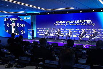 Podiumsdiskussion auf einer internationalen Konferenz der GSIS: Mehrere Personen sitzen auf einer Bühne vor einem großen blauen Bildschirm mit der Aufschrift ‚World Order Disrupted: Implications for innovation and security‘. Links ist ein Redner an einem Podium zu sehen, während im Hintergrund und auf einer großen Projektionsfläche sein Bild angezeigt wird. Im Vordergrund sind Zuhörer in einem Konferenzsaal erkennbar.
