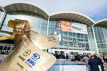 Großes Schiffspropeller-Modell vor dem Eingang der Hamburg Messe. Im Hintergrund die verglaste Fassade des Messegebäudes mit SMM-Banner und zahlreichen Besucherinnen und Besuchern vor dem Haupteingang.