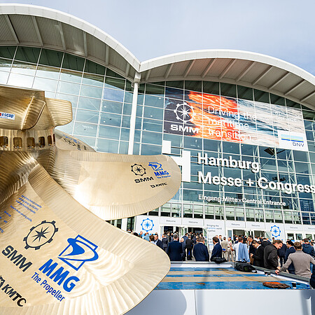 Großes Schiffspropeller-Modell vor dem Eingang der Hamburg Messe. Im Hintergrund die verglaste Fassade des Messegebäudes mit SMM-Banner und zahlreichen Besucherinnen und Besuchern vor dem Haupteingang.