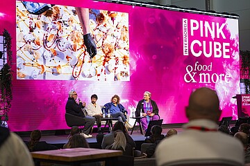 Auf einer Bühne vor einem großen Publikum sitzen vier Personen in Sesseln und führen eine Podiumsdiskussion. Hinter ihnen erstreckt sich eine sehr große, leuchtend pinke LED‑Wand mit dem Schriftzug „INTERNORGA PINK CUBE & food more“. Links auf der Wand ist ein großes Foto eingeblendet, das eine kulinarische Kreation mit verschiedenen Zutaten und farbenfrohen Saucen zeigt. Die Bühne ist mit Mikrofonen und Wassergläsern ausgestattet. Im Vordergrund sind die Silhouetten einiger Zuschauerinnen und Zuschauer zu sehen, die der Diskussion folgen.