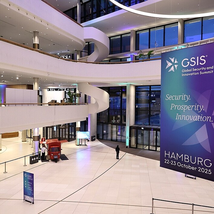 Große, moderne Eingangshalle eines Konferenzgebäudes mit offenen Galerieebenen und hoher Glasfront. Ein großes Banner kündigt den „GSIS – Global Security and Innovation Summit“ in Hamburg für den 22.–23. Oktober 2025 an.