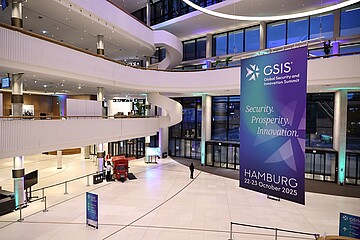 Große, moderne Eingangshalle eines Konferenzgebäudes mit offenen Galerieebenen und hoher Glasfront. Ein großes Banner kündigt den „GSIS – Global Security and Innovation Summit“ in Hamburg für den 22.–23. Oktober 2025 an.