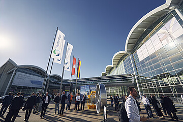 Besucherinnen und Besucher vor den Messehallen der Hamburg Messe während der WindEnergy Hamburg. Vor dem Gebäude stehen Fahnen der Veranstaltung und eine technische Ausstellungskomponente im Außenbereich.