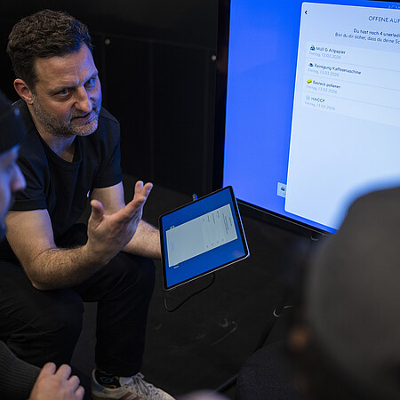 Drei Personen besprechen die digitale Verwaltung offener Aufgaben. Auf einem großen Bildschirm und einem Tablet ist eine Checkliste mit noch zu erledigenden Aufgaben zu sehen.