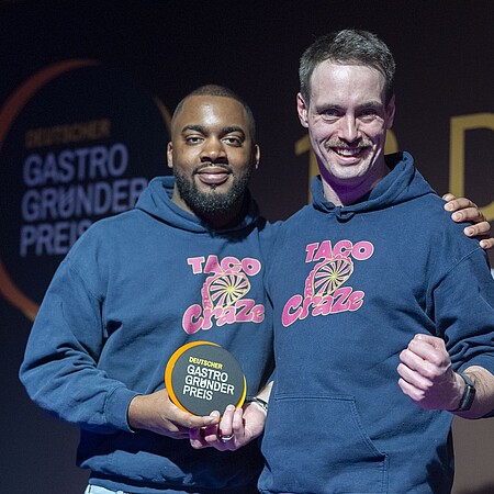 Zwei Personen in blauen „TACO Craze“-Hoodies stehen nebeneinander und halten eine runde Auszeichnung mit der Aufschrift „DEUTSCHER GASTRO GRÜNDER PREIS“.