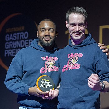Zwei Personen in blauen „TACO Craze“-Hoodies stehen nebeneinander und halten eine runde Auszeichnung mit der Aufschrift „DEUTSCHER GASTRO GRÜNDER PREIS“.