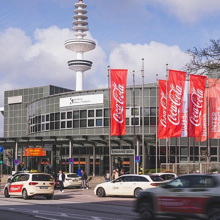 Eine Straßenansicht vor einem Messeeingang mit mehreren vorbeifahrenden Autos. Am Gebäude hängen rote und weiße Fahnen mit Marken- und Messebeschriftung, im Hintergrund steht ein hoher Turm.