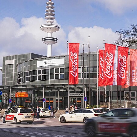 Eine Straßenansicht vor einem Messeeingang mit mehreren vorbeifahrenden Autos. Am Gebäude hängen rote und weiße Fahnen mit Marken- und Messebeschriftung, im Hintergrund steht ein hoher Turm.
