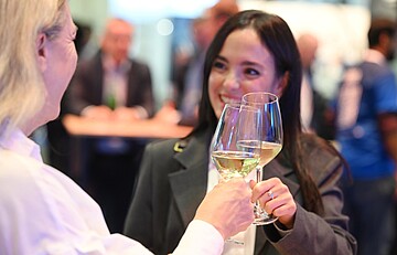 Zwei Personen stoßen auf einer Veranstaltung mit Gläsern Weißwein an. Im Hintergrund stehen weitere Menschen an hohen Tischen in einer hell beleuchteten Umgebung.