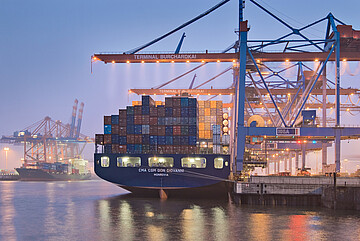 Containerschiff am Terminal Burchardkai im Hamburger Hafen bei Abenddämmerung, umgeben von großen Containerkränen.
