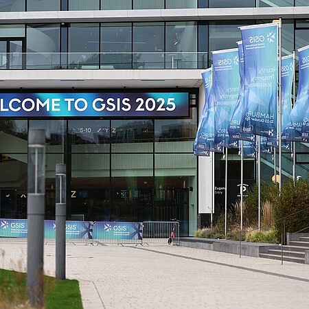 Eingangsbereich des modernen Congress Center Hamburg (CCH) mit einem großen digitalen Banner, auf dem ‚Welcome to GSIS 2025‘ steht. Rechts auf dem Vorplatz wehen mehrere blau‑türkise GSIS-Flaggen.