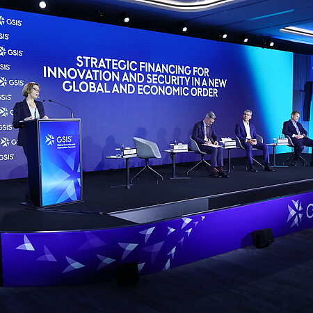 Konferenzbühne beim GSIS (Global Security and Innovation Summit) in Hamburg. Eine Person spricht an einem Rednerpult vor einer großen blauen LED-Wand mit der Aufschrift „Strategic Financing for Innovation and Security in a New Global and Economic Order“. Mehrere Teilnehmende sitzen auf der Bühne in einer Podiumsdiskussion. Rechts zeigt ein großer Bildschirm die sprechende Person. Die Veranstaltung findet in einem modern beleuchteten Konferenzsaal statt.