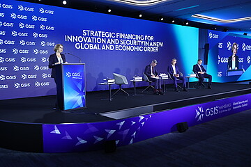 Konferenzbühne beim GSIS (Global Security and Innovation Summit) in Hamburg. Eine Person spricht an einem Rednerpult vor einer großen blauen LED-Wand mit der Aufschrift „Strategic Financing for Innovation and Security in a New Global and Economic Order“. Mehrere Teilnehmende sitzen auf der Bühne in einer Podiumsdiskussion. Rechts zeigt ein großer Bildschirm die sprechende Person. Die Veranstaltung findet in einem modern beleuchteten Konferenzsaal statt.