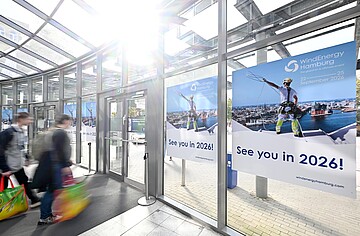 Heller Eingangsbereich eines Messegeländes mit großen Glasflächen. Mehrere Besucher gehen durch die Eingangshalle. An den Fenstern hängen großformatige Plakate der WindEnergy Hamburg mit dem Hinweis „See you in 2026!“ und den Veranstaltungsdaten 22.–25. September 2026.