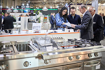 Messestand mit professionellen Küchengeräten, darunter ein großer Edelstahlherd mit mehreren Gasbrennern und Spülbecken im Vordergrund. Im Hintergrund stehen mehrere Personen zusammen und betrachten Unterlagen, während weitere Geräte und Displays sichtbar sind.