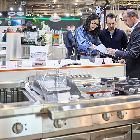 Messestand mit professionellen Küchengeräten, darunter ein großer Edelstahlherd mit mehreren Gasbrennern und Spülbecken im Vordergrund. Im Hintergrund stehen mehrere Personen zusammen und betrachten Unterlagen, während weitere Geräte und Displays sichtbar sind.