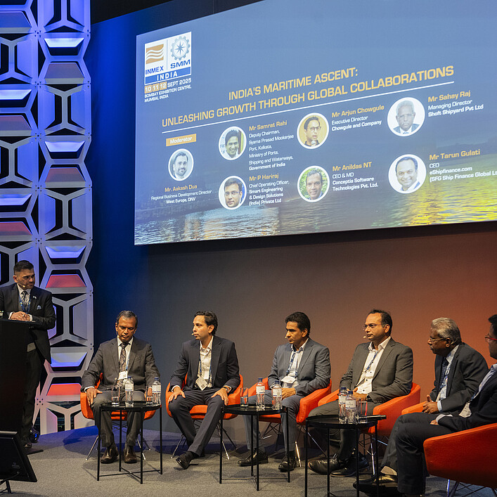 Podiumsdiskussion auf einer Konferenzbühne. Eine Person steht an einem Rednerpult mit der Aufschrift „Open Stage“, während sechs weitere Personen auf orangefarbenen Sesseln sitzen. Im Hintergrund zeigt eine große Leinwand Informationen zu einer maritimen Fachveranstaltung in Indien. Die Bühne ist in Blau- und Orangetönen beleuchtet und von dekorativen Wandpaneelen eingerahmt.