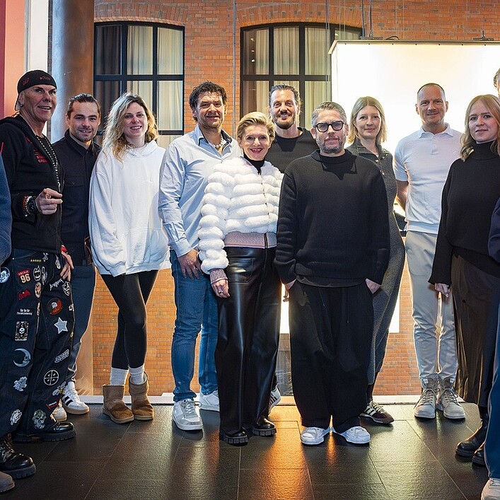 Eine Gruppe von Menschen steht für ein gemeinsames Foto in einem Innenraum vor einer Backsteinwand. Einige Personen stehen, andere sitzen, und die Gruppe trägt überwiegend legere Kleidung. Helle Studiobeleuchtung befindet sich hinter ihnen und sorgt für eine freundliche, ausgeleuchtete Szene.