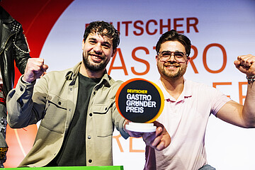 Deutscher Gastro-Gründerpreis: Zwei Personen präsentieren auf einer Bühne den Pokal des Deutschen Gastro-Gründerpreises und halten die Fäuste nach oben.