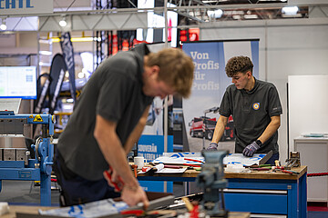 Zwei Personen arbeiten an einer Werkbank in einer Messe- oder Ausstellungshalle. Auf dem Tisch liegen Werkzeuge und bearbeitete Blechteile. Im Vordergrund ist eine Person unscharf bei der Metallbearbeitung zu sehen, während eine weitere Person an einem Werkstück arbeitet. Im Hintergrund befinden sich Maschinen, Messestände und ein Banner mit der Aufschrift „Von Profis für Profis“.