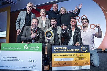 Eine Gruppe von neun Personen posiert auf einer Bühne vor einem großen orange‑weißen Hintergrund mit der Aufschrift „Deutscher Gastro‑Gründerpreis“. Vor der Gruppe stehen zwei große symbolische Schecks: links ein grüner Scheck über 3.000 Euro für einen Ausbildungsplatz bei Simmet‑Training, rechts ein orangefarbener Hauptgewinn‑Scheck über 10.000 Euro mit dem Gewinnernamen „MOI“. Davor steht zudem eine große Flasche Sekt. Eine Person in der Mitte hält eine runde Auszeichnungstrophäe hoch. Die Gruppe zeigt teils jubelnde Gesten und wirkt wie bei einer Siegerehrung. Geben Sie Ihr Feedback zu BizChat ab.